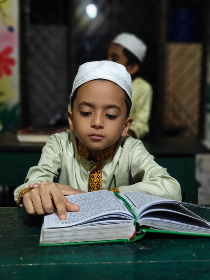 Services-02 A focused young boy in traditional attire reads a religious text inside a classroom.